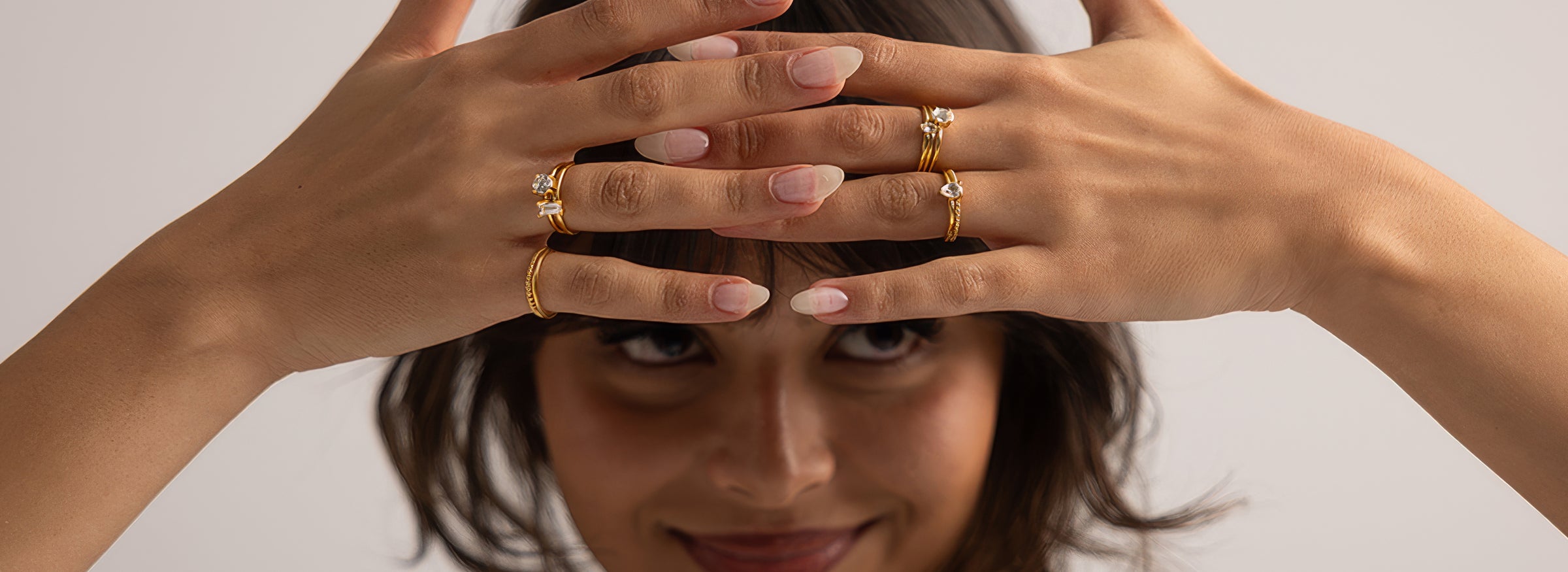 A close-up lifestyle shot of a woman with her hands raised to her forehead, showcasing several gold rings with clear gemstones on her fingers against a soft, light gray background.