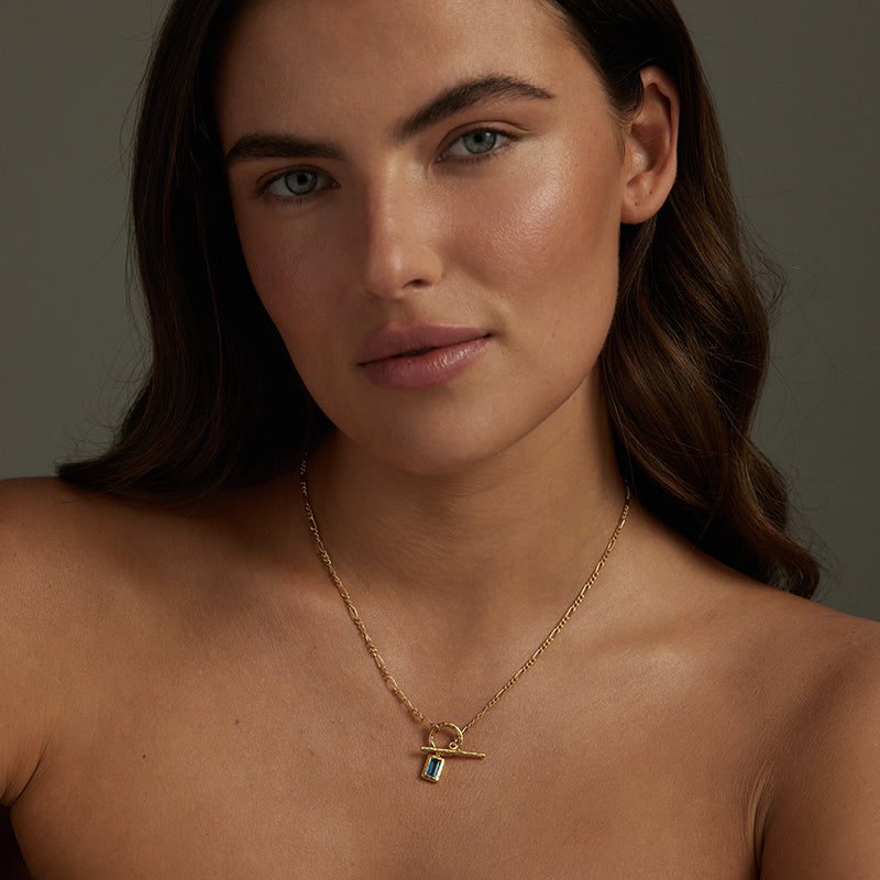A close-up of a woman wearing a delicate gold chain necklace with a small pendant featuring a blue rectangular stone, set against a dark gray background.