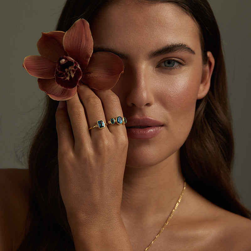 A close-up lifestyle shot of a model wearing gold rings with blue stones, holding a brown orchid flower near her eye against a dark gray background.