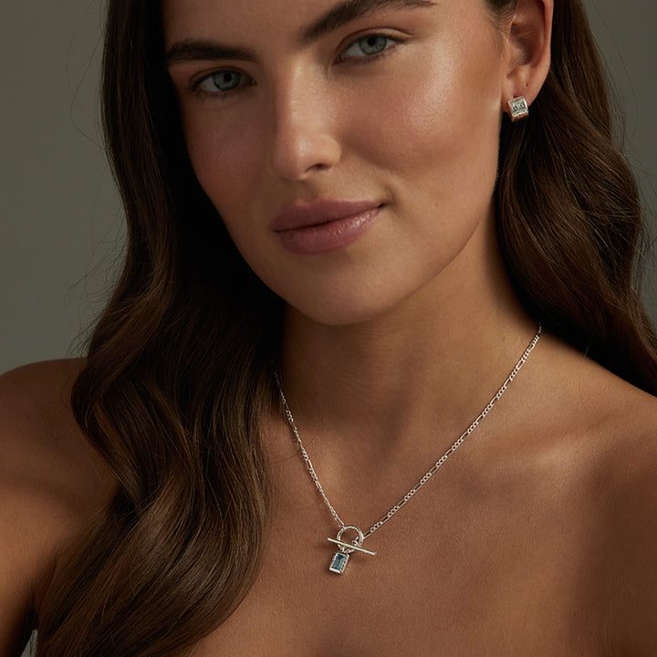 A close-up of a woman wearing a delicate silver chain necklace with a small pendant featuring a light blue rectangular stone, along with a matching silver stud earring.