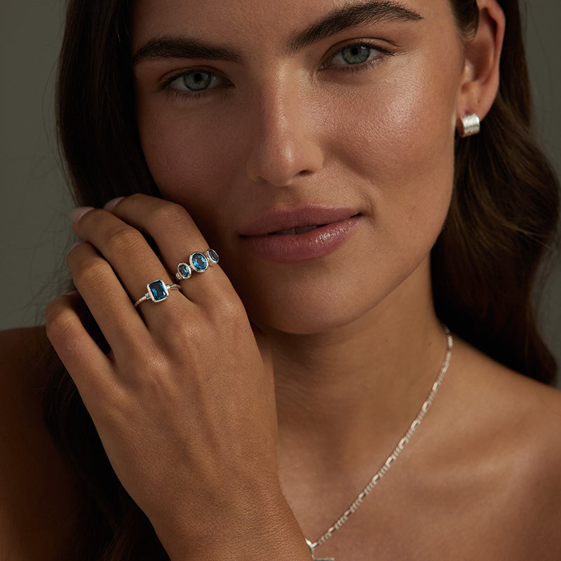 A close-up lifestyle shot of a woman wearing silver rings with blue gemstones, a silver stud earring, and a delicate silver chain necklace.