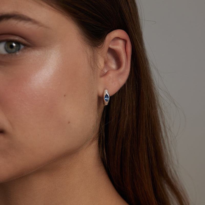 A close-up of a woman's ear wearing a small marquise-shaped stud earring with a deep blue center and a silver-toned halo setting.