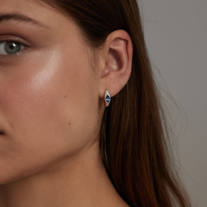 A close-up of a woman's ear wearing a small marquise-shaped stud earring with a deep blue center and a silver-toned halo setting.
