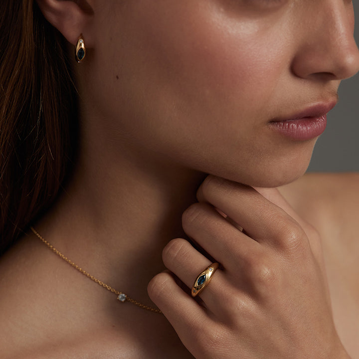 Close-up of a woman wearing a gold ring with a dark blue oval stone on her finger, matching gold drop earrings, and a delicate gold chain necklace.