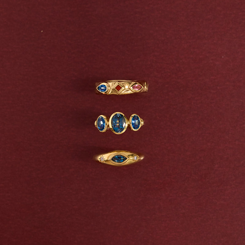 Three gold rings with blue and red gemstones arranged vertically on a dark red background.
