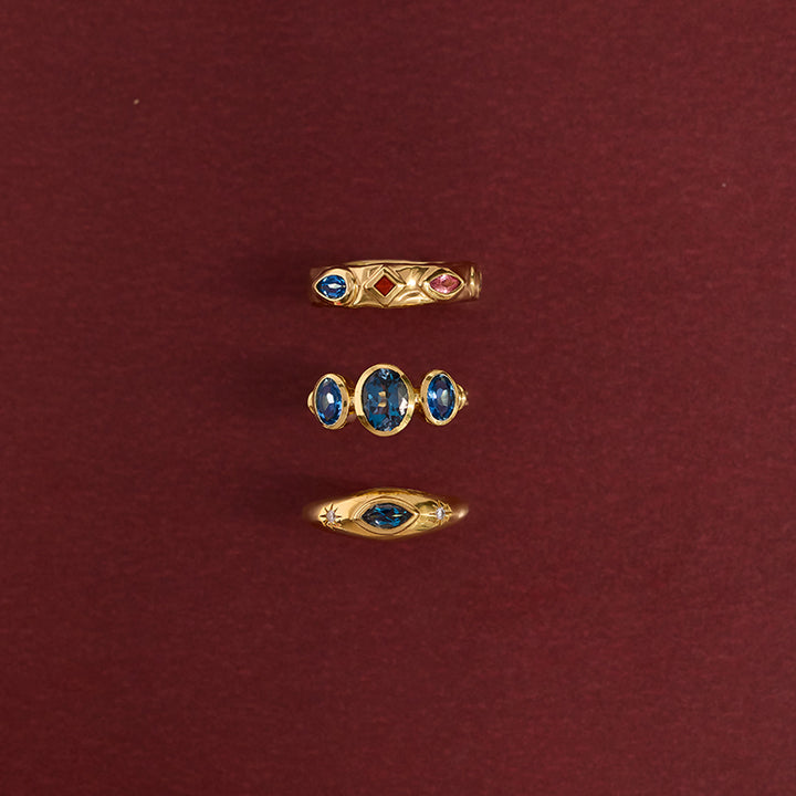 Three gold rings with blue and red gemstones arranged vertically on a dark red background.