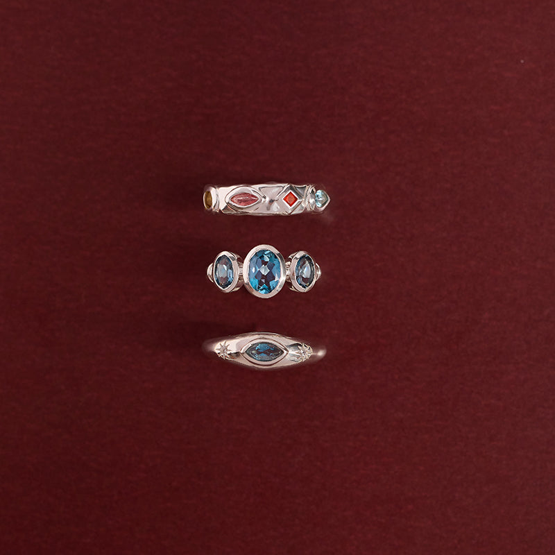 Three silver rings with blue and red gemstones arranged vertically on a dark red background.