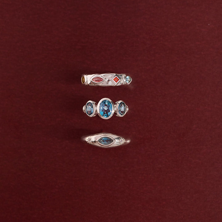 Three silver rings with blue and red gemstones arranged vertically on a dark red background.