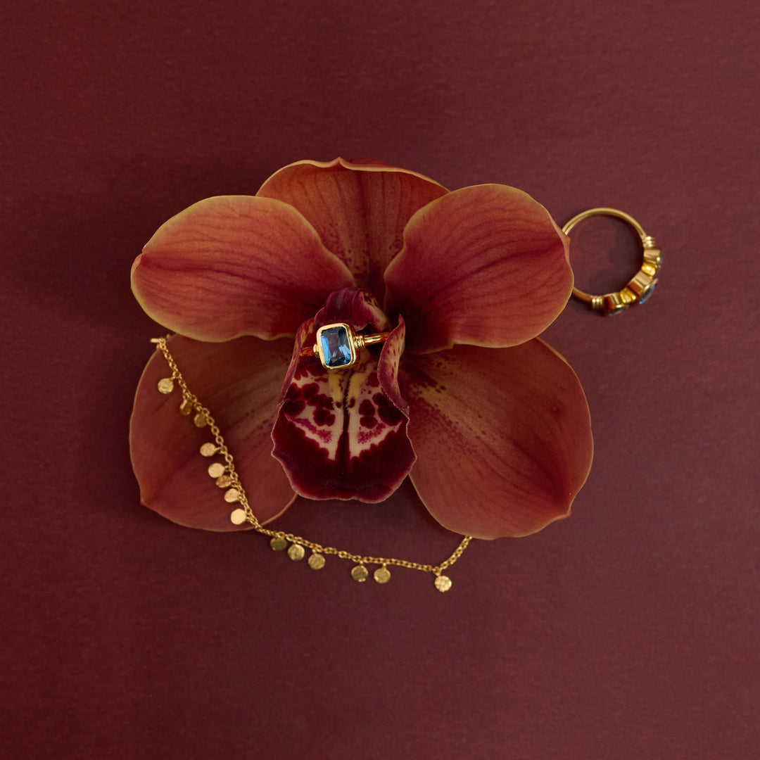 A gold ring with a square blue gemstone is displayed on a large maroon orchid flower, with a delicate gold coin chain draped underneath and a textured gold ring resting on the right side, all set against a solid dark maroon background.