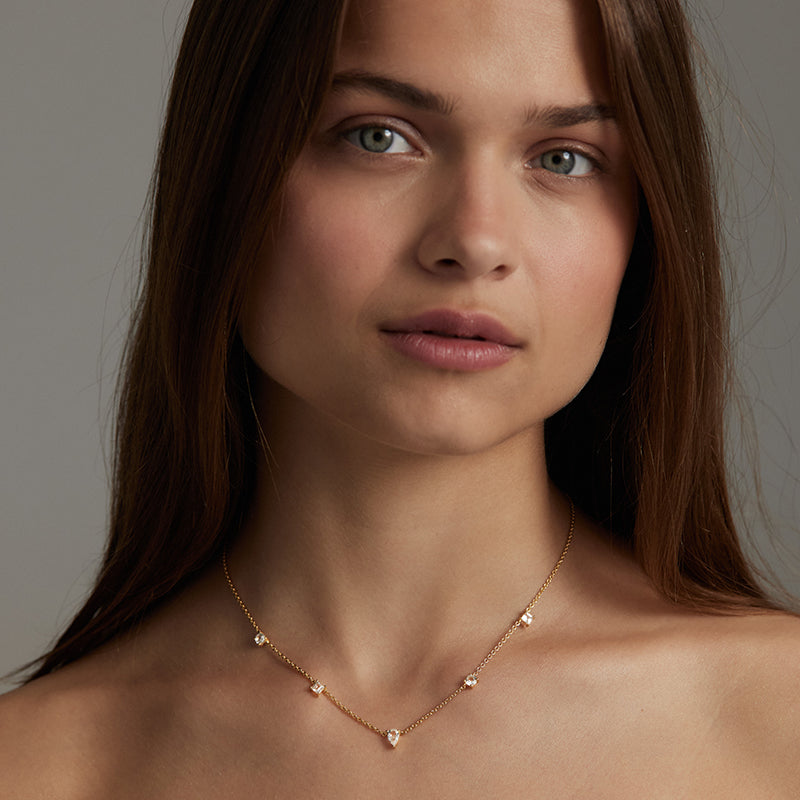 A close-up portrait of a woman wearing a delicate rose gold necklace with small star-shaped clear gemstones against a neutral gray background.