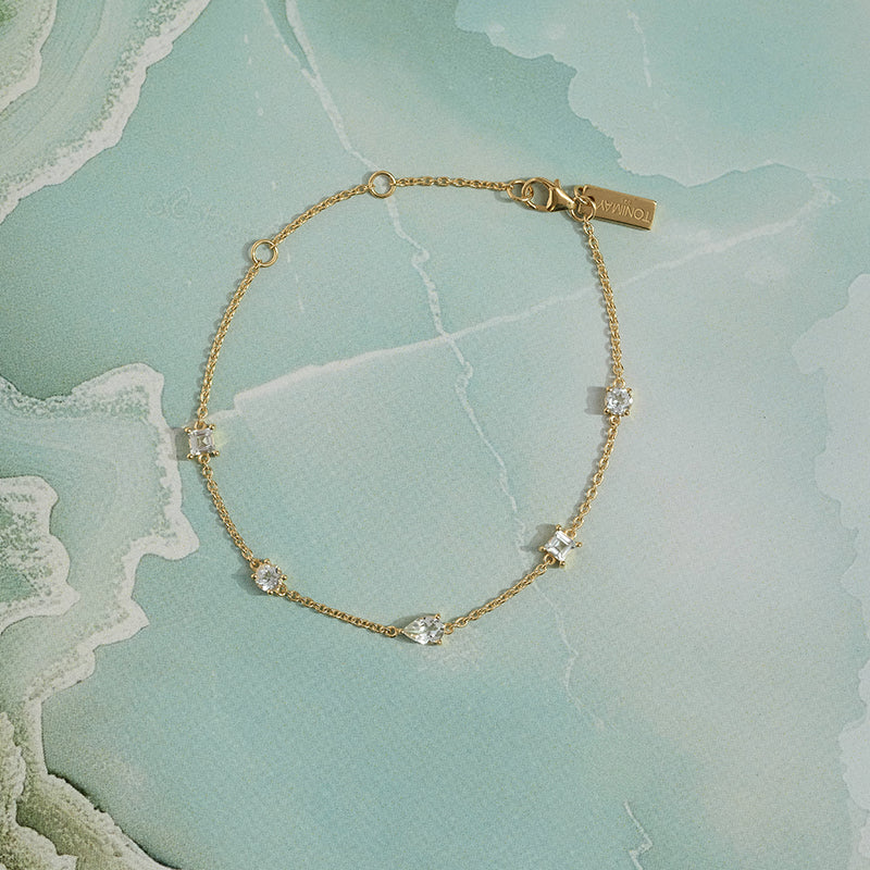 A delicate gold chain bracelet with small clear gemstone accents, displayed on a light blue-green marbled surface. The brand name 'ToniMay' is engraved on the clasp tag.
