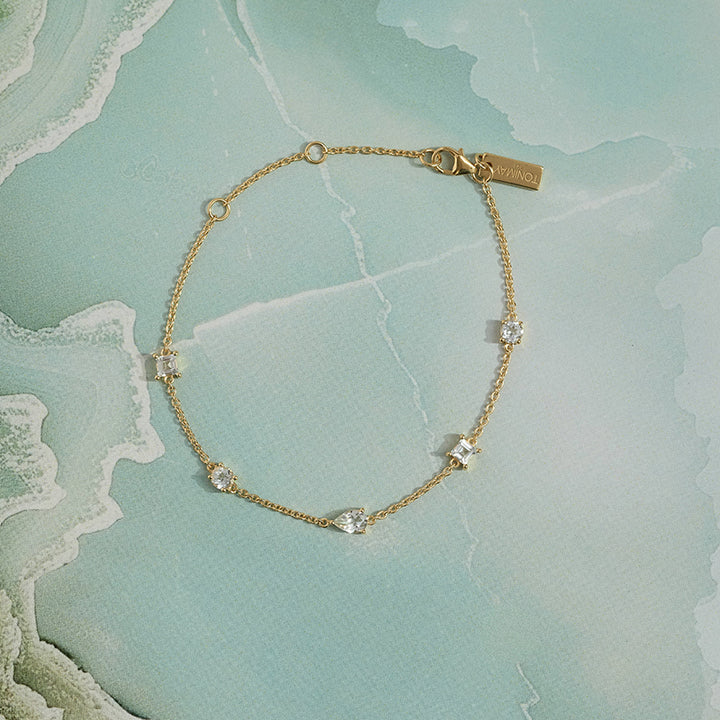 A delicate gold chain bracelet with small clear gemstone accents, displayed on a light blue-green marbled surface. The brand name 'ToniMay' is engraved on the clasp tag.