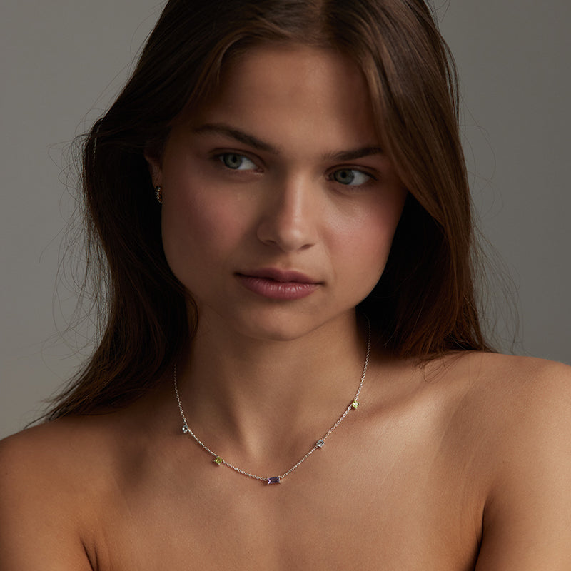 A close-up portrait of a woman wearing a delicate silver-toned necklace with small multicolored gemstone accents, set against a neutral gray background.