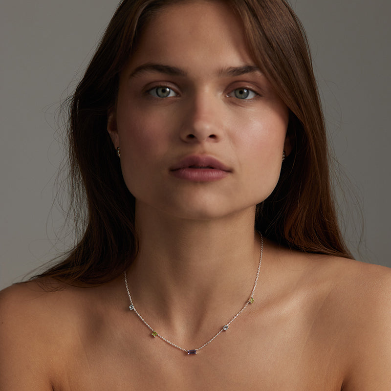 A close-up portrait of a woman wearing a delicate silver-toned necklace with small multicolored gemstone accents, set against a neutral gray background.