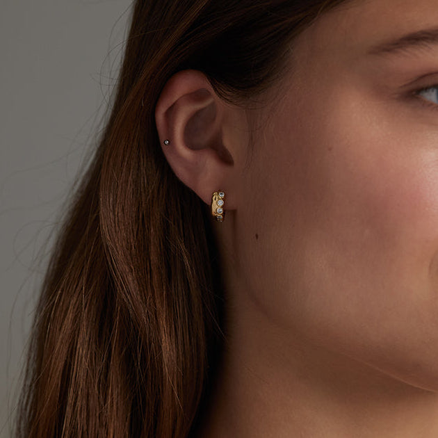 Close-up of a woman's ear wearing a small gold hoop earring with clear stones, against a soft gray background.