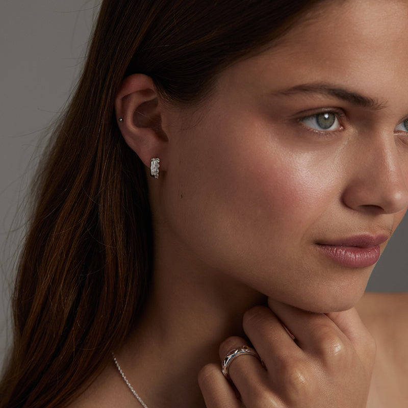 A close-up portrait of a woman wearing a small silver hoop earring and a silver ring, with her hand near her neck against a soft gray background.