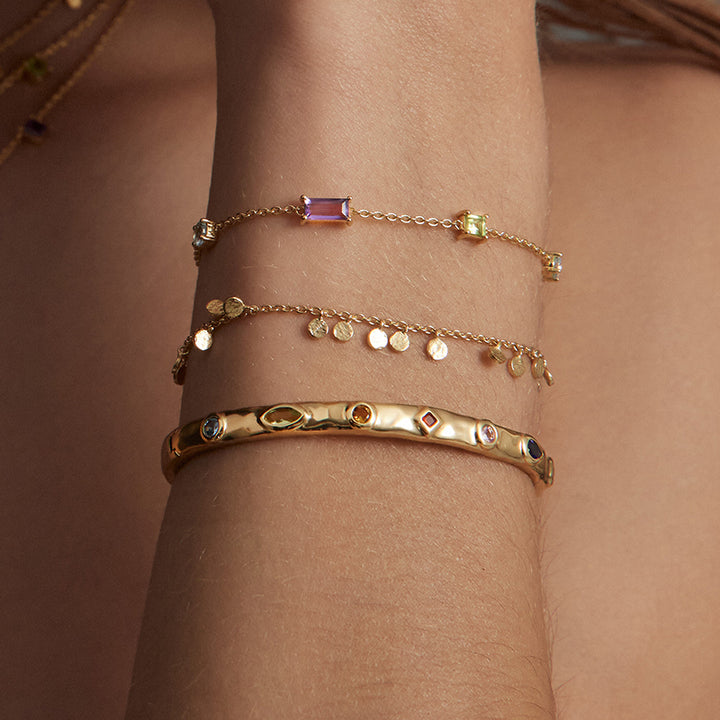 Three gold-toned bracelets stacked on a person's wrist, including a thin chain bracelet with a rectangular pink gemstone, a charm bracelet with small round gold discs, and a thicker bangle with oval and square inset stones.