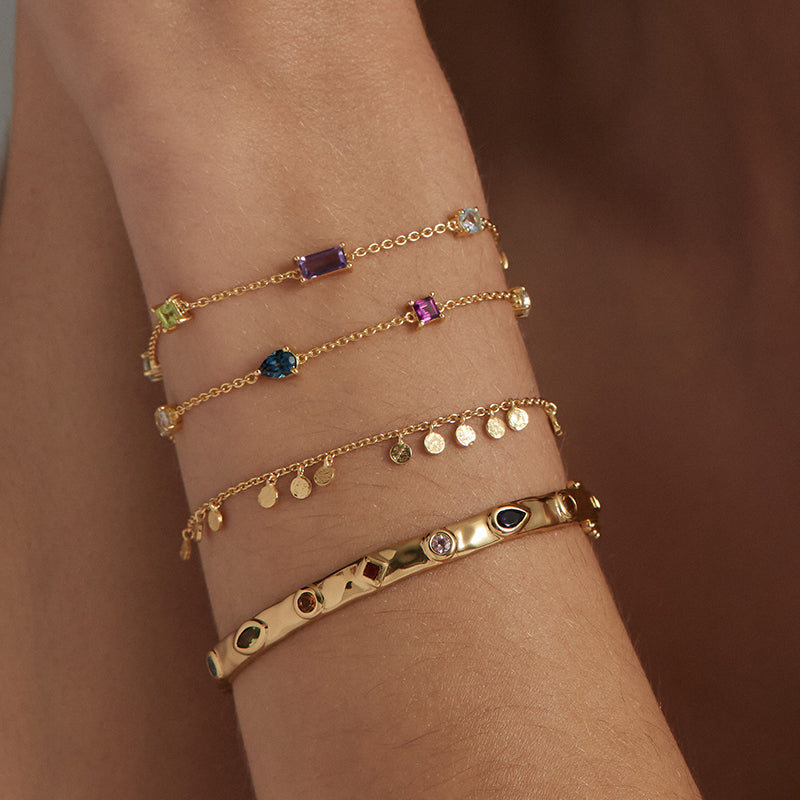 Four delicate gold chain bracelets with various colorful gemstone and charm details, stacked on a person's wrist against a warm brown background.