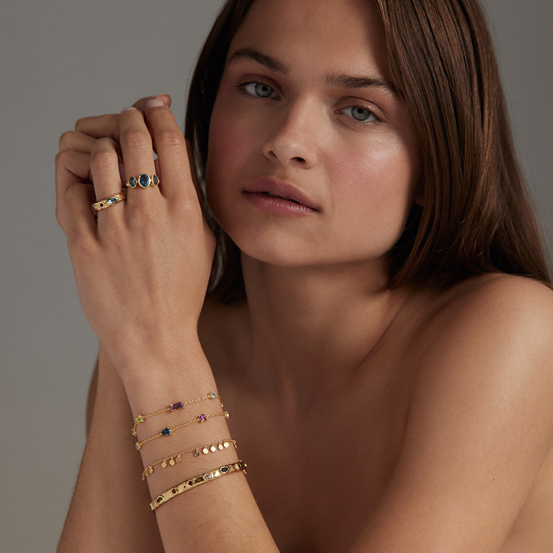A close-up lifestyle shot of a woman wearing delicate gold bracelets and rings on her wrist and fingers, set against a neutral gray background.