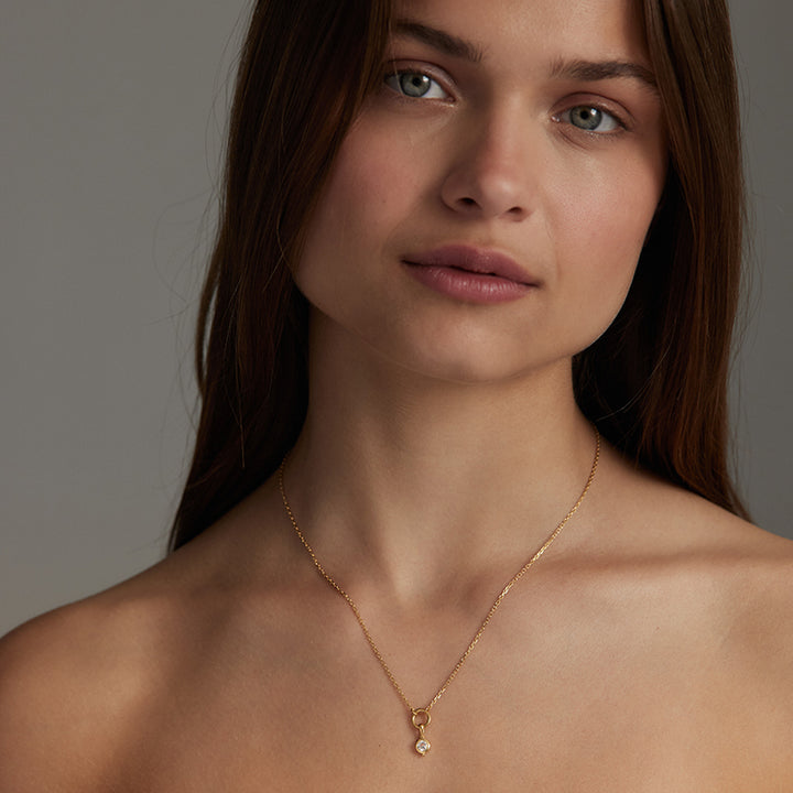 A close-up of a woman wearing a delicate gold chain necklace with a small white pearl pendant, set against a neutral gray background.