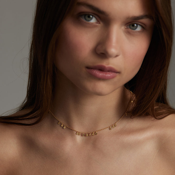 A close-up of a woman wearing a delicate gold chain necklace with small, irregular gold charms, set against a neutral gray background.