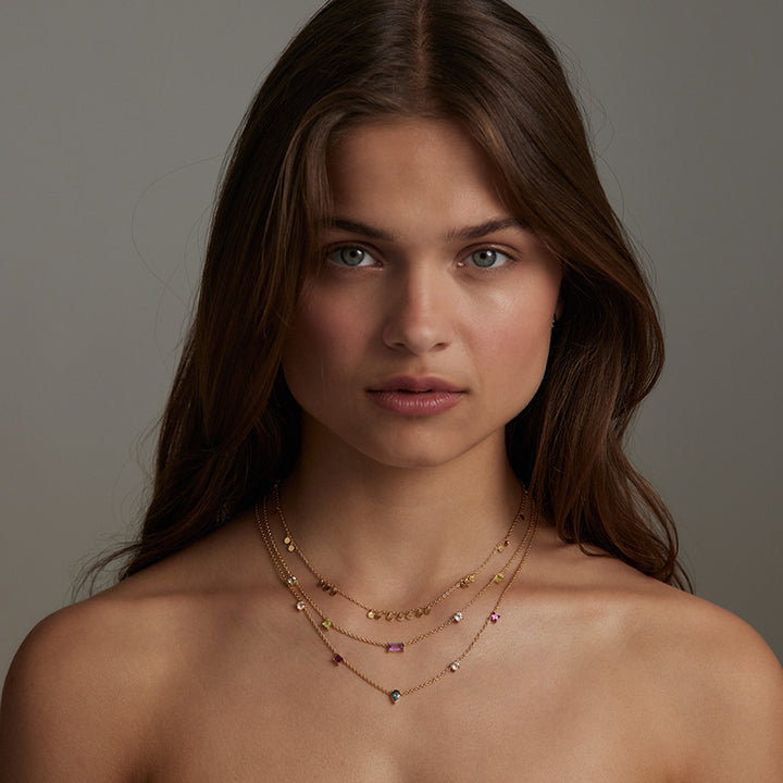 A close-up portrait of a woman with long brown hair wearing delicate layered gold necklaces with small colorful gemstone accents against a neutral gray background.