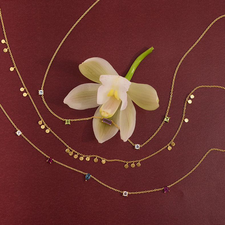 Gold chain necklaces with small geometric and dangling charms, arranged on a dark burgundy background with a pale orchid flower at the center.