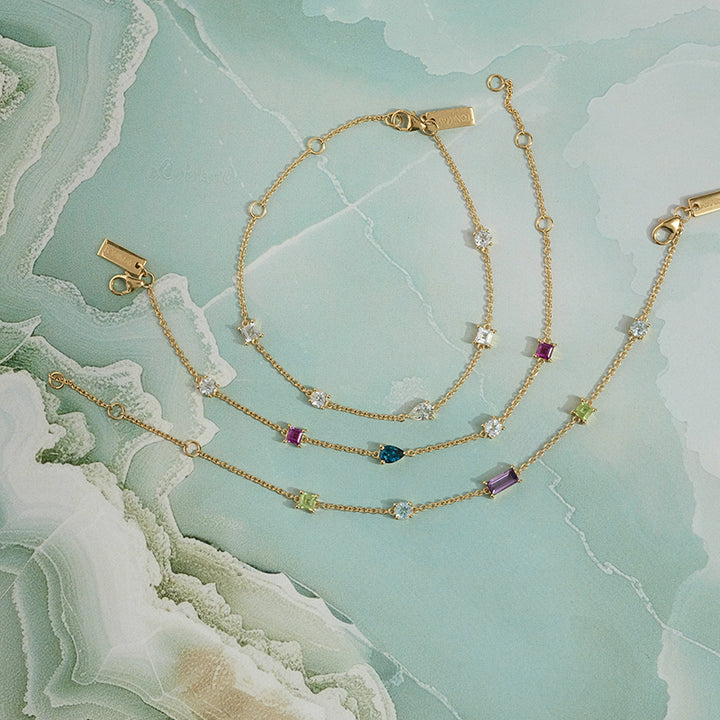 Three delicate gold chain bracelets with small star-shaped settings and colorful gemstones, arranged on a light aqua marbled surface.
