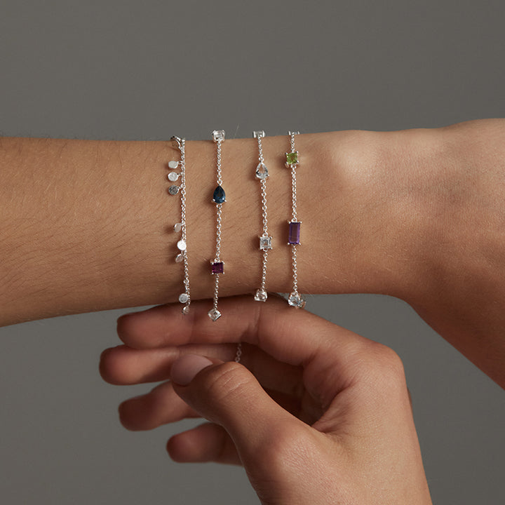 Four delicate silver chain bracelets with colorful gemstones are shown worn on a person's wrist, with the other hand gently touching them against a plain gray background.
