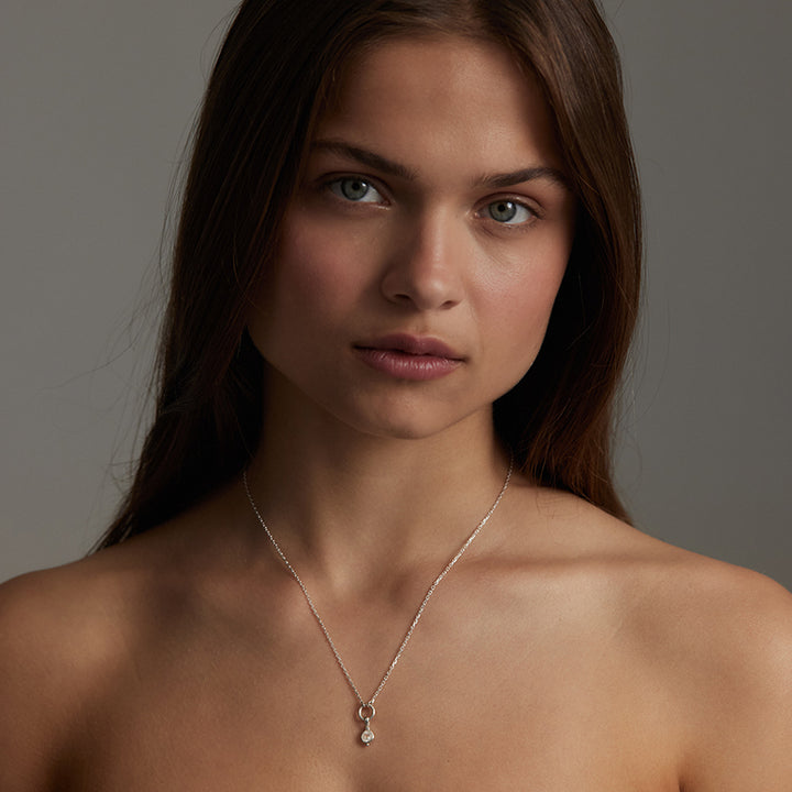 A close-up portrait of a woman wearing a delicate silver chain necklace with a small silver heart-shaped pendant, set against a neutral gray background.