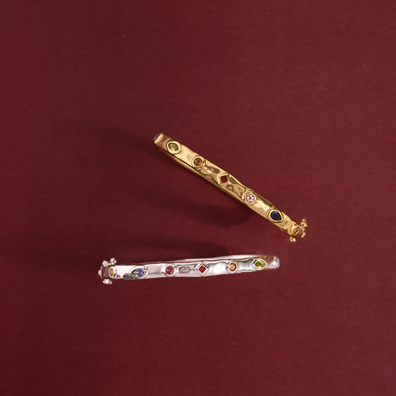 Two slim bangles, one gold-toned and one silver-toned, both decorated with small multicolored stones, displayed on a solid dark red background.