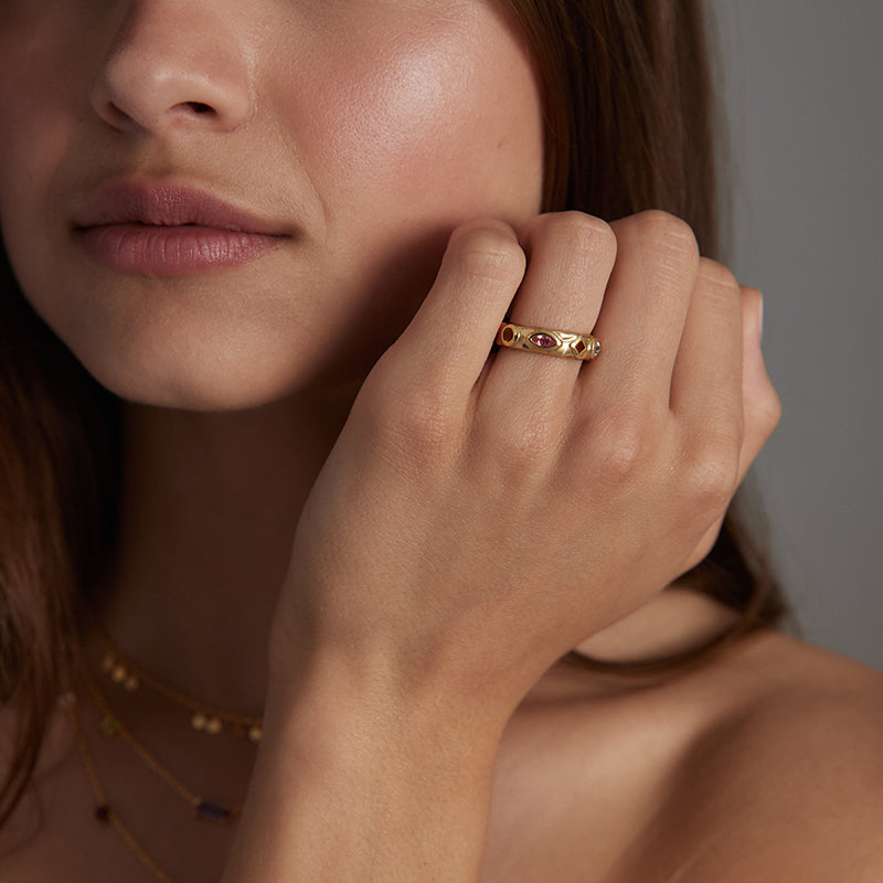 A close-up lifestyle shot of a model wearing a delicate gold band ring with small red gemstone accents on her finger.