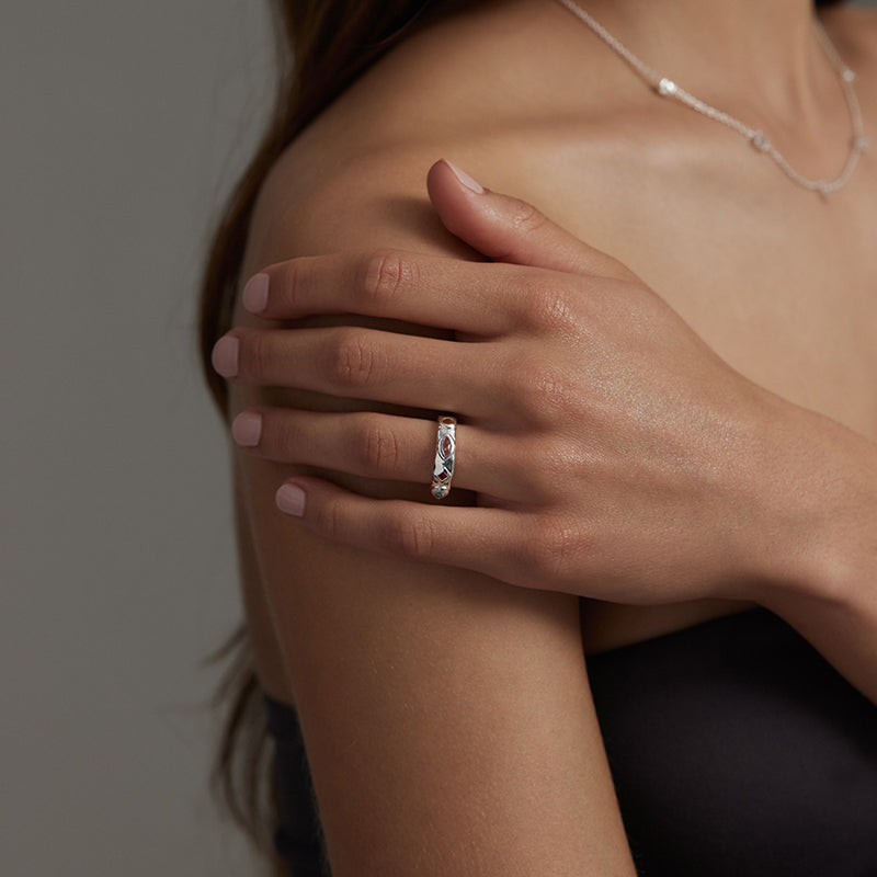 A close-up lifestyle shot of a woman's hand resting on her shoulder, wearing a slim silver band ring set with clear stones. The background is a soft, neutral gray.