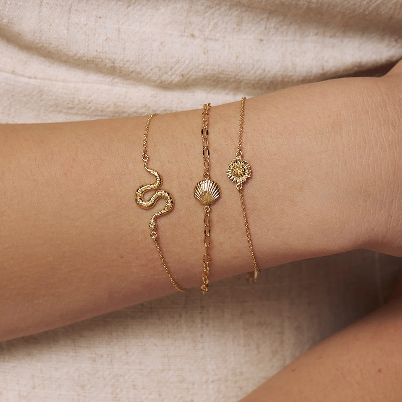 Three delicate gold bracelets with snake, shell, and sun motifs worn on a person's wrist against a soft beige fabric background.