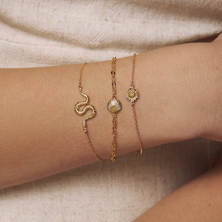 Three delicate gold bracelets with snake, shell, and sun motifs worn on a person's wrist against a soft beige fabric background.