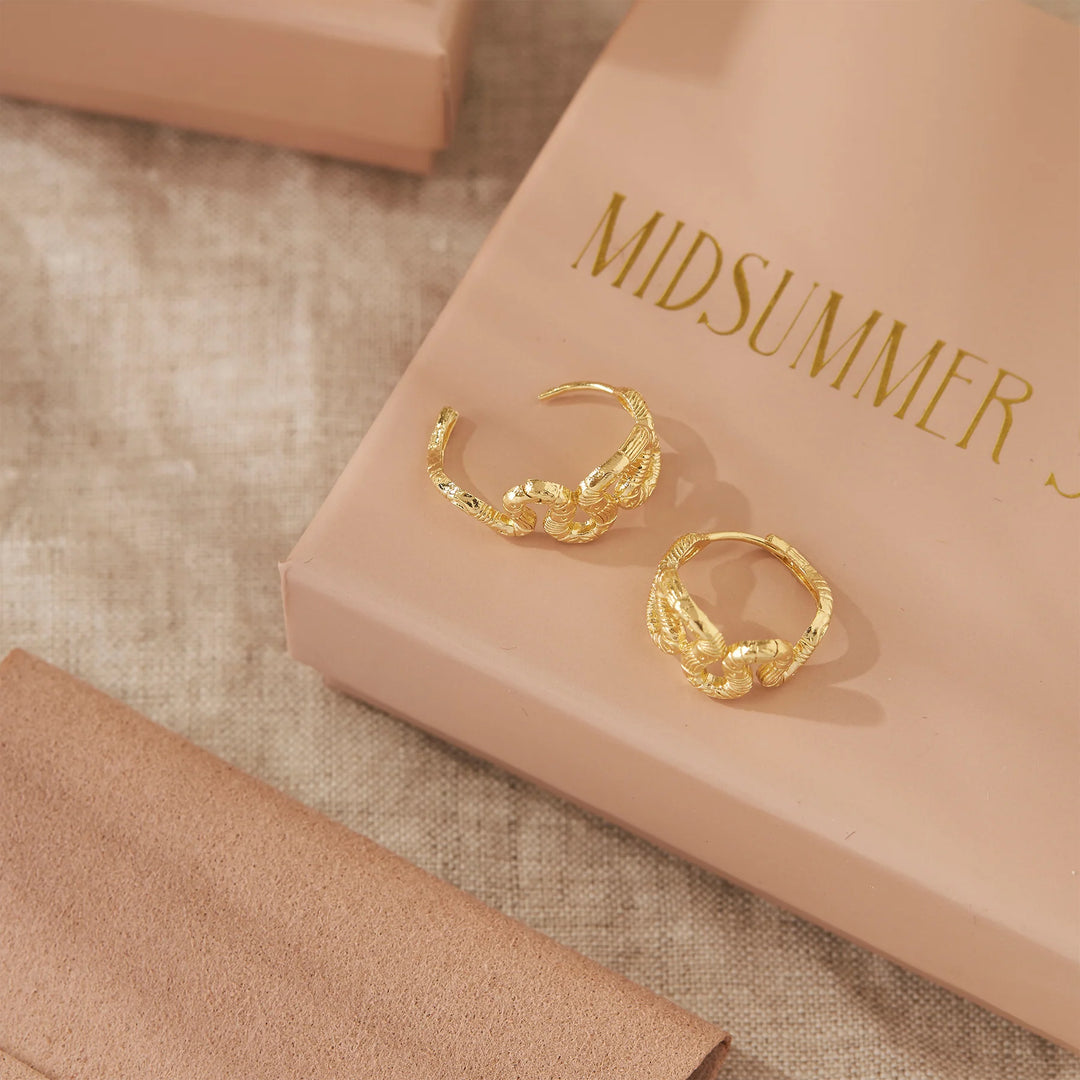 A pair of textured gold hoop earrings displayed on a beige box with the brand name 'MIDSUMMER' printed in gold lettering. The box is set on a neutral beige fabric background.