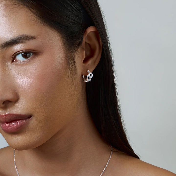 A close-up of a woman wearing a small silver earring with a stylized interlocking loop design, shown against a plain light gray background.