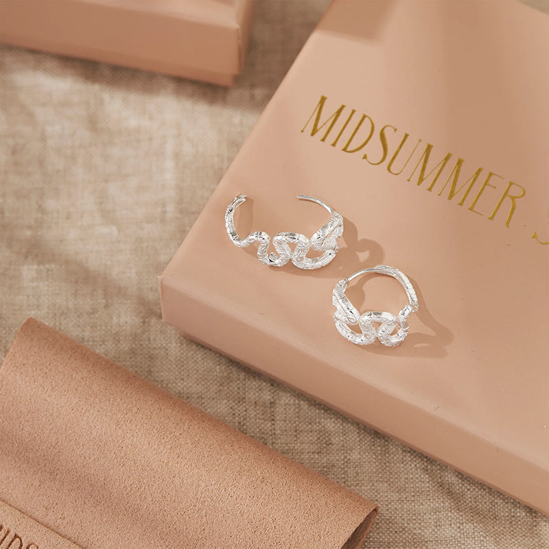 A pair of silver huggie hoop earrings with a textured, leaf-like or snake-like sculpted design, displayed on a beige box with gold lettering that reads 'MIDSUMMER'.