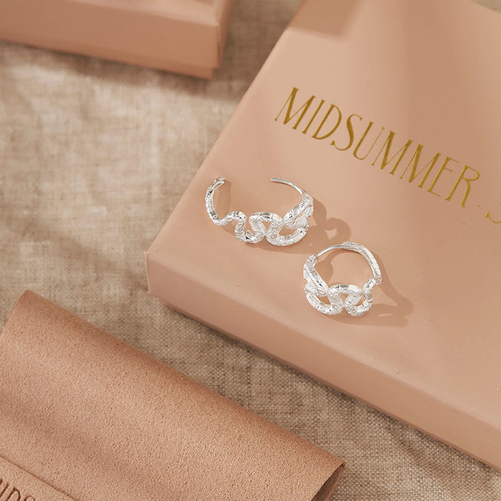 A pair of silver huggie hoop earrings with a textured, leaf-like or snake-like sculpted design, displayed on a beige box with gold lettering that reads 'MIDSUMMER'.