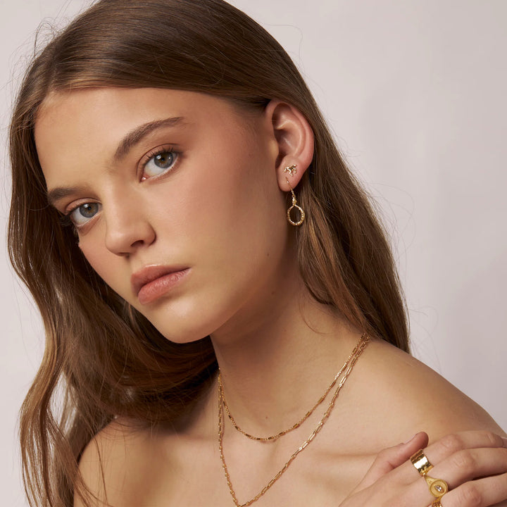 A close-up portrait of a woman wearing delicate gold jewelry, including a small hoop earring, layered gold necklaces, and a ring, against a soft light gray background.