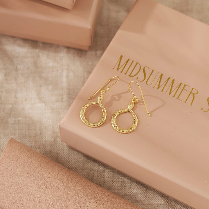 A pair of gold-tone drop earrings with textured circular hoops and fishhook wires, displayed on a peach-colored jewelry box with the brand name 'MIDSUMMER S.