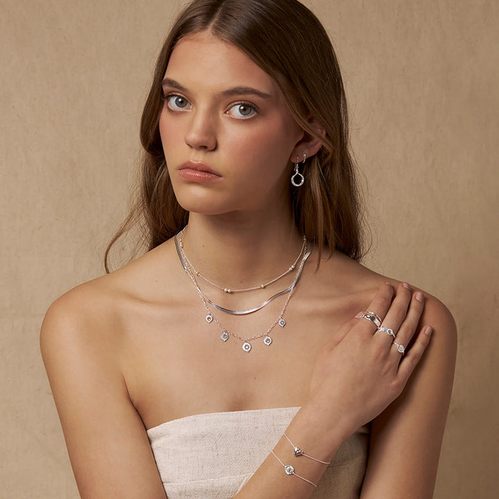 A woman models a delicate layered silver jewelry set including a necklace with small circular charms, a matching ring, and an earring against a warm beige textured background.