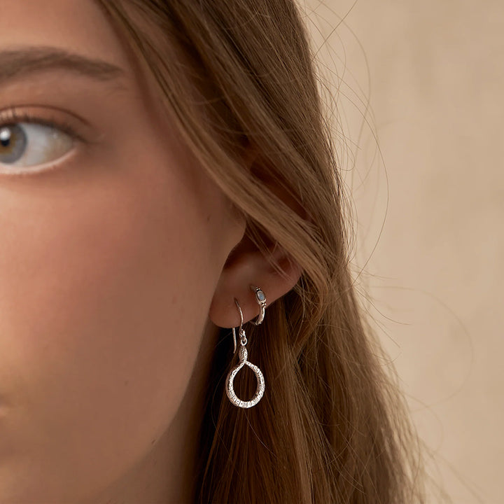 A close-up of a woman's ear wearing a silver huggie hoop earring with a dangling textured silver ring earring, against a soft beige background.
