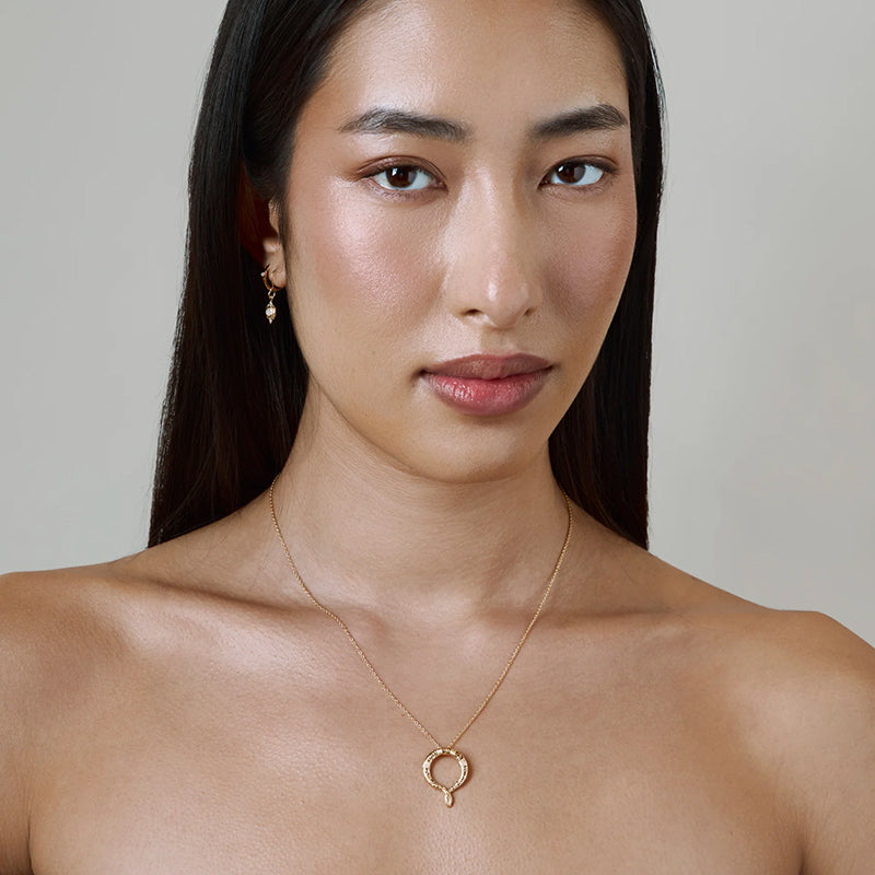 A close-up of a woman wearing a delicate gold necklace with a small open heart pendant and a matching gold hoop earring, set against a plain light gray background.