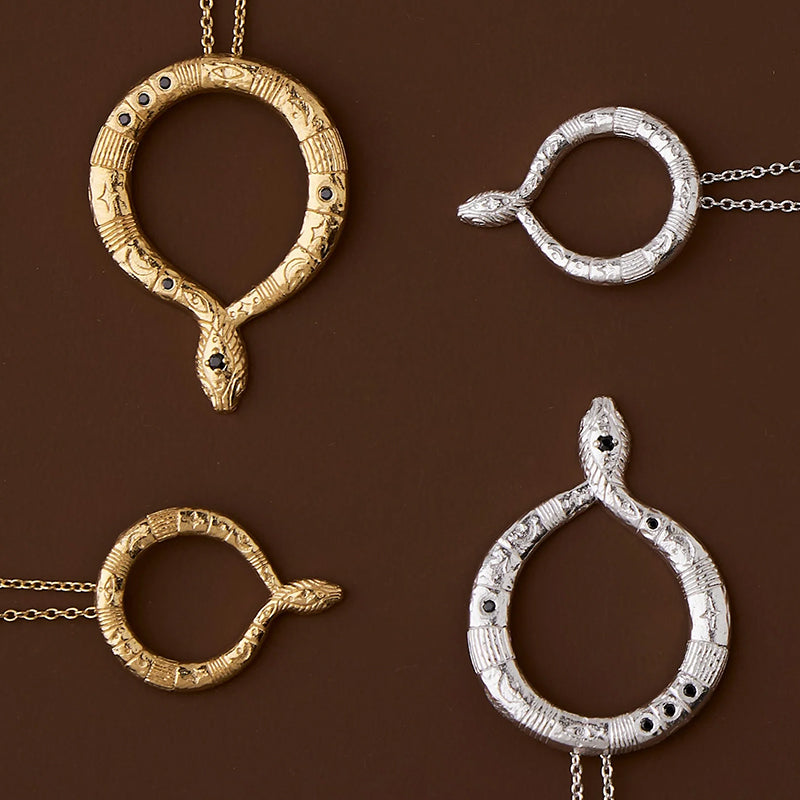 Four snake pendant necklaces arranged on a dark brown background, featuring oxidized silver and yellow gold finishes with black stone eyes and engraved details.