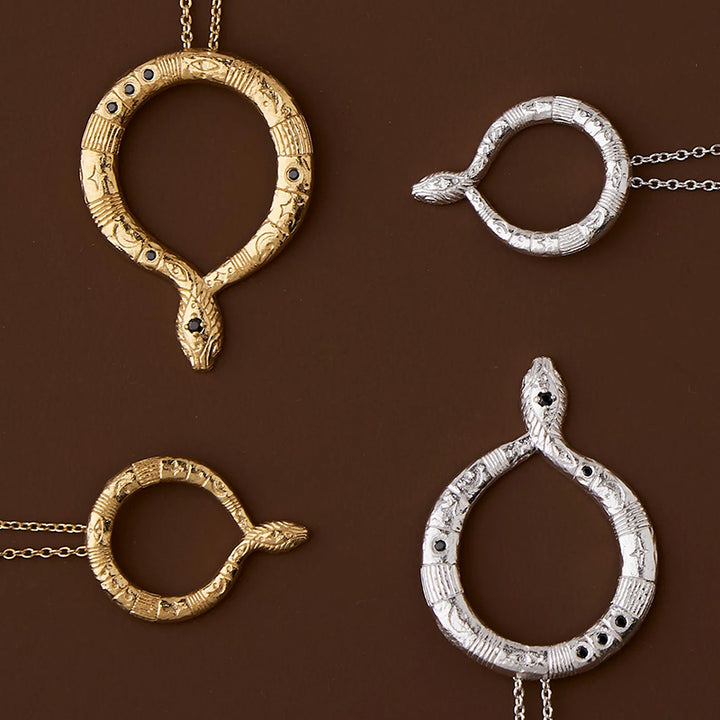 Four snake pendant necklaces arranged on a dark brown background, featuring oxidized silver and yellow gold finishes with black stone eyes and engraved details.
