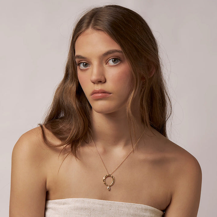 A woman with long brown hair wears a delicate gold necklace with a small circular pendant, photographed against a soft light gray background.